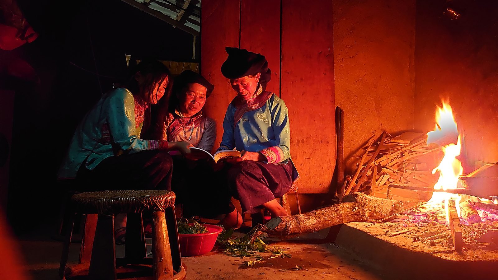 Ba người phụ nữ ngồi quanh bếp lửa trong nhà, ánh lửa hắt lên vách gỗ